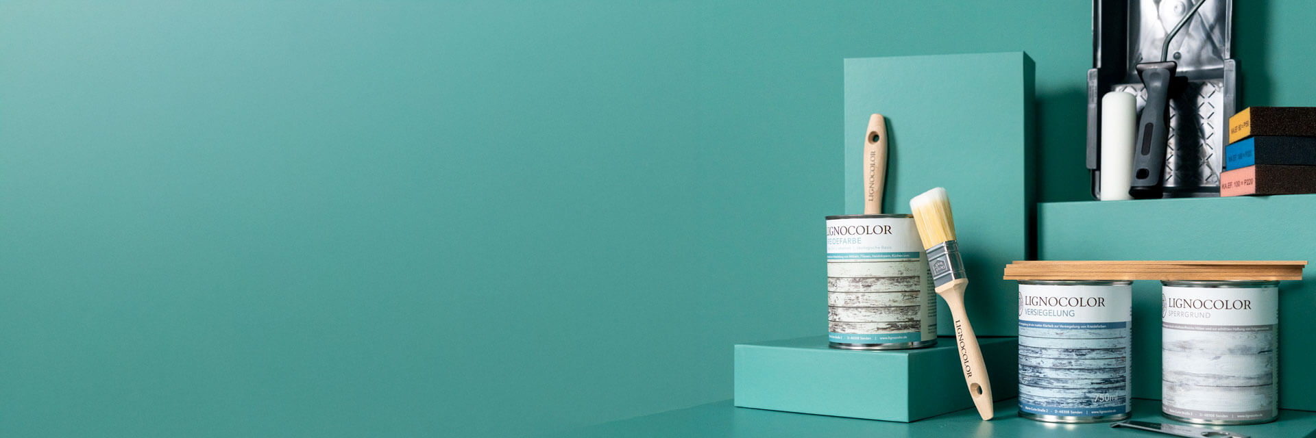 Mehrere Farbdosen mit Pinsel, Farbwanne, Schleifschwämmen und Farbroller vor einer grünen Wand