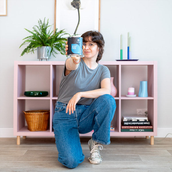 Frau sitzt vor einem IKEA Kallax und hält Buntlack Dose in die Kamera