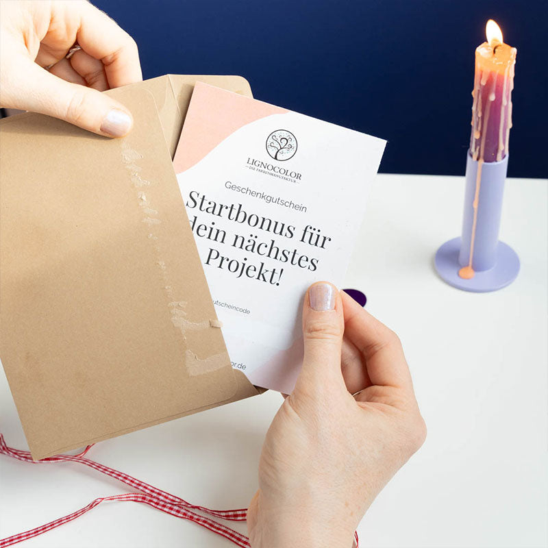 Gutscheinkarte auf einem weißen Tisch mit einer Kerze vor blauer Wand wird in einen braunen Umschlag eingepackt