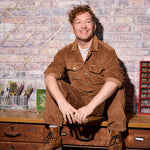 Thomas Klotz in einem braunen Arbeitsoutfit sitzend auf einer Werkbank